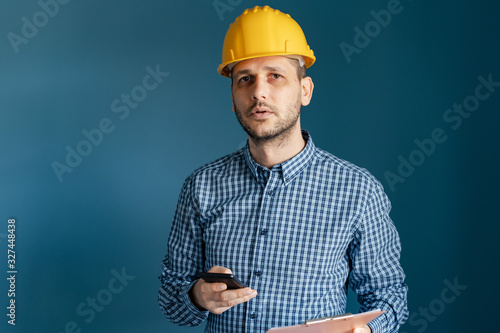 Portrait of young engineer or building contractor wearing yellow protective helmet and shirt standing in front of blue wall background holding smart phone and clipboard looking to camera