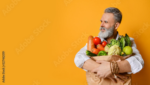 Cool old mature senior man with gray beard shopping hold grocery shopping bag with healthy organic vegetables on yellow