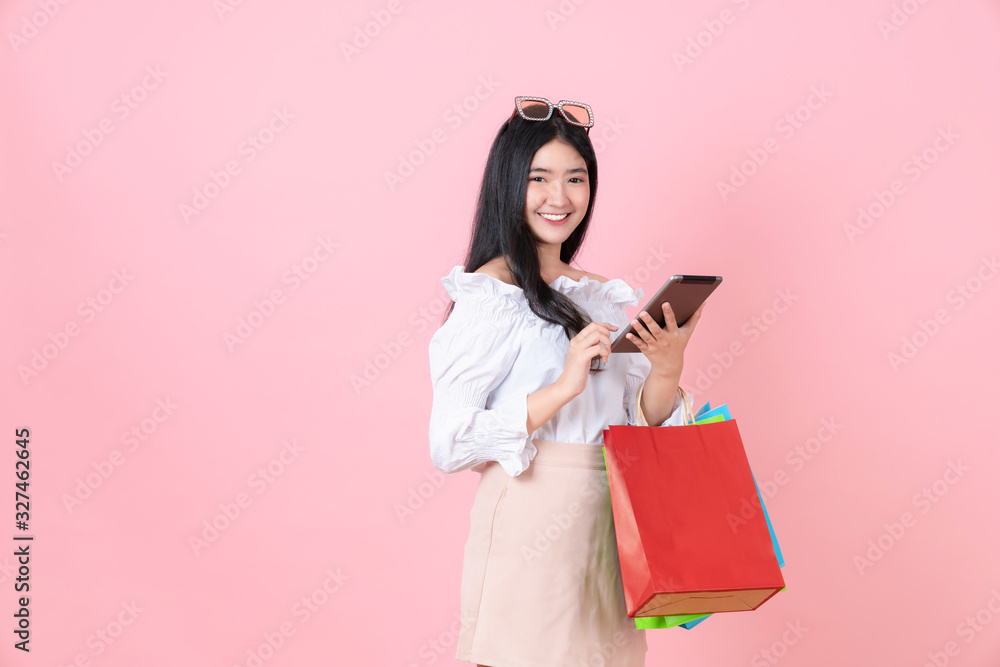 Cheerful beautiful Asian woman holding multi coloured shopping bags and digital tablet on light pink background. Cheerful beautiful Asian woman holding multi coloured shopping bags and digital tablet on light pink background.