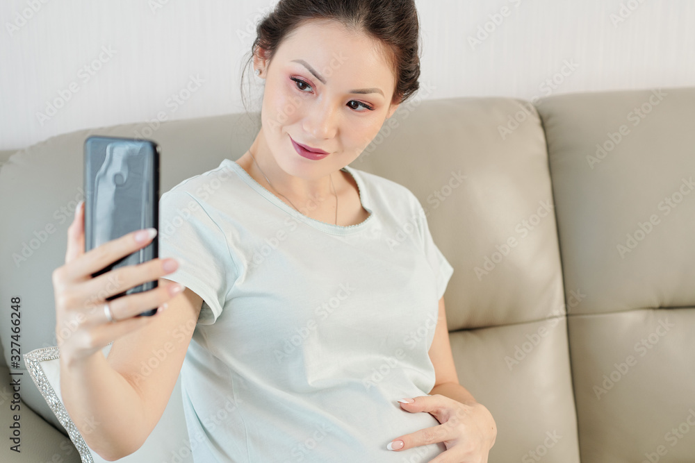 Lovely smiling young Asian woman sitting on sofa and taking selfie for social media Lovely smiling young Asian woman sitting on sofa and taking selfie for social media