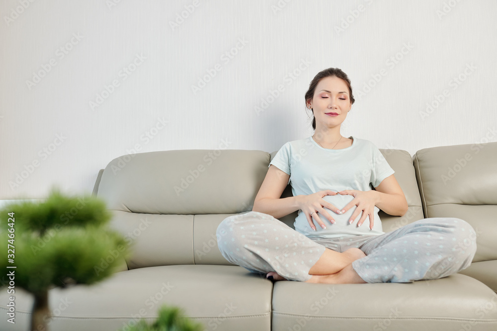 Beautiful smiling young Vietnamese woman touching her belly when meditating on sofa at home Beautiful smiling young Vietnamese woman touching her belly when meditating on sofa at home