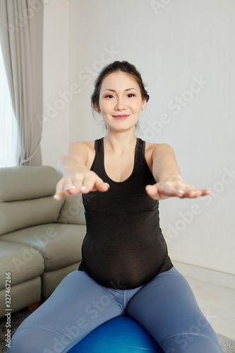 Smiling happy young pregnant woman outstretching arms when doing exercise on yoga ball at home