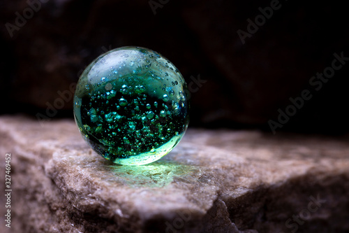 Close up of a vibrant teal green transparent glass marble shining its light on the reflecting textured surface of a white stone with dark background photo