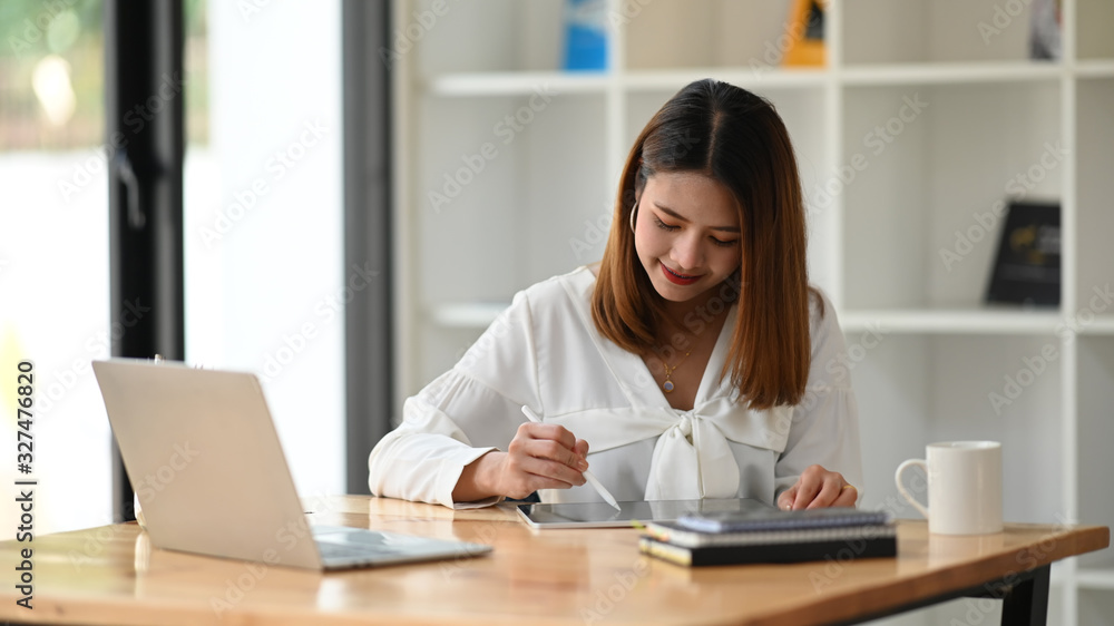 Woman holding stylus pen and using it to drawing on her computer tablet while sitting in front her laptop at the modern working table with office glass wall as background. Woman holding stylus pen and using it to drawing on her computer tablet while sitting in front her laptop at the modern working table with office glass wall as background.