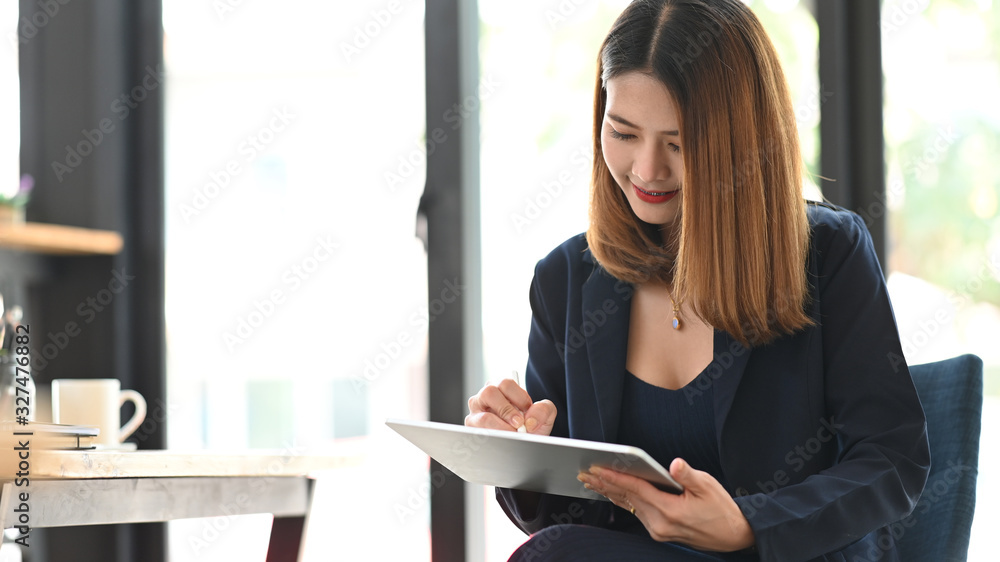 Executive woman holding a computer tablet and stylus pen in her hands while sitting at the modern working table with office glass wall as background. Executive woman holding a computer tablet and stylus pen in her hands while sitting at the modern working table with office glass wall as background.