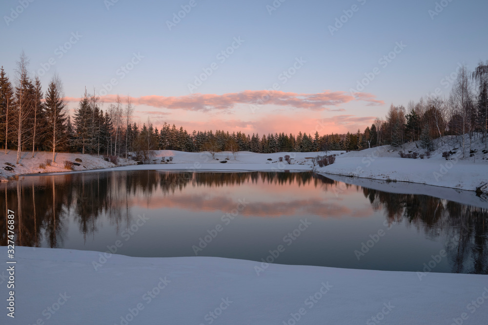 Winter landscape. The forest lake is partially covered with ice, surrounded by rocks, shrubs and spruces against the evening sky painted by the rays of the setting sun.