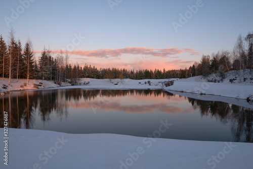 Winter landscape. The forest lake is partially covered with ice, surrounded by rocks, shrubs and spruces against the evening sky painted by the rays of the setting sun. © Александр Овсянников