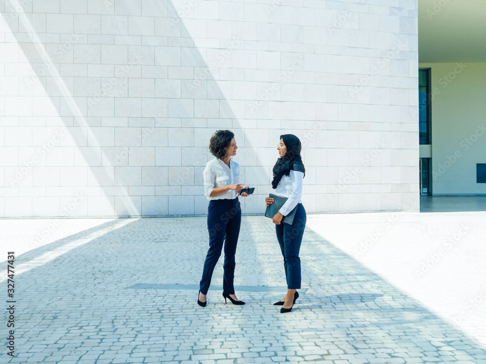 © Mangostar - Multiethnic female business colleagues discussing report outside. Women in office suits and hijab standing outdoors, talking and gesturing. Multicultural business team concept