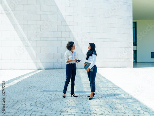 Multiethnic female business colleagues discussing report outside. Women in office suits and hijab standing outdoors, talking and gesturing. Multicultural business team concept photo