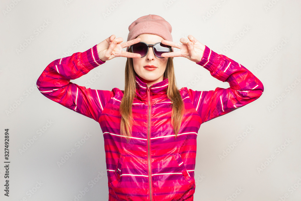 Young woman with glasses, a hat and pink sports jacket makes cool hand gesture, holds her hands in her face, white background with copy space. Concept modern trendy style. Face expression. Banner Young woman with glasses, a hat and pink sports jacket makes cool hand gesture, holds her hands in her face, white background with copy space. Concept modern trendy style. Face expression. Banner