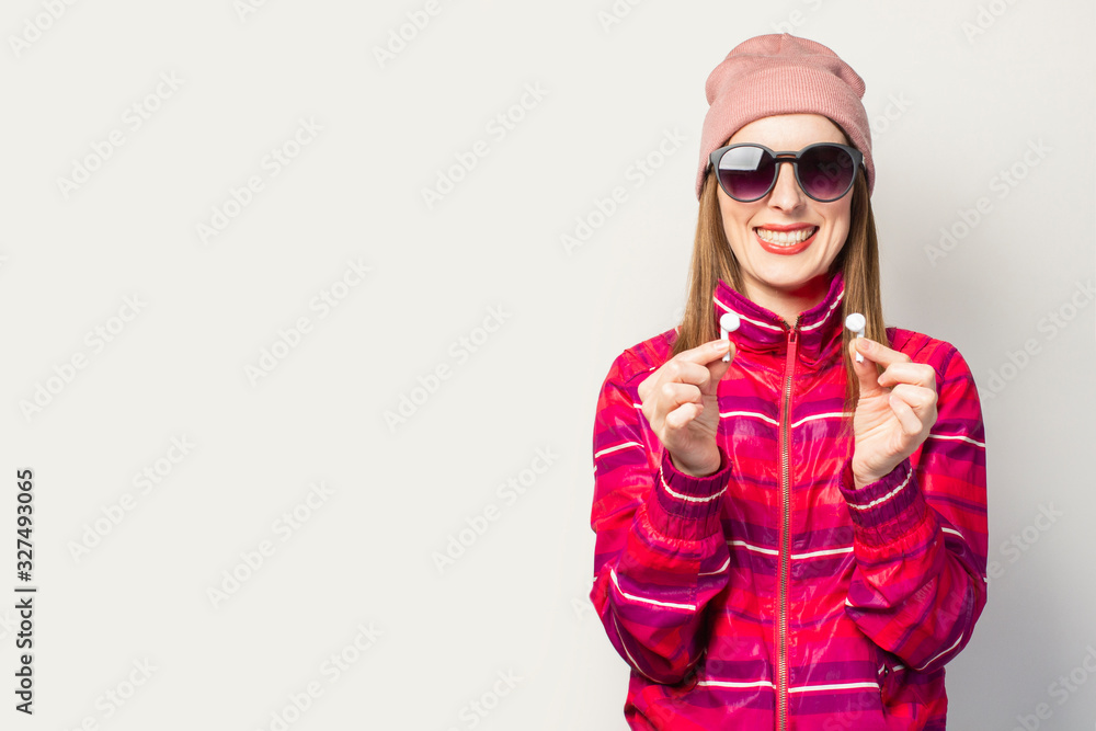 Friendly Young woman in glasses, hat and pink sports jacket with a smiley face holds wireless headphones on a white background. Concept modern style, cool music. Face expression. Banner