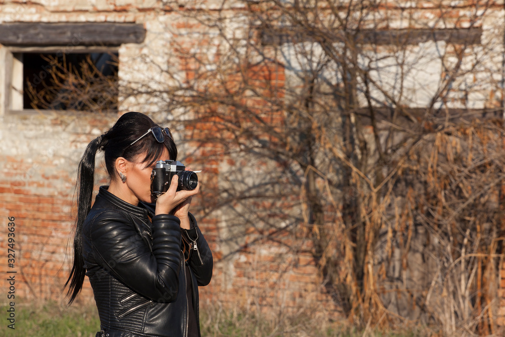 beautiful girl in a black leather jacket poses next to an old collapsed building, holds a cigarette and old camera in her hands