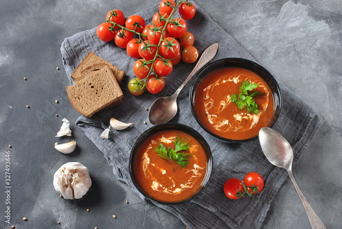 Hot winter soup. Red hot tomato soup with garlic, sweet paprika, parsley, served with cream and sourdough bread in two black ceramic bowls on a gray background