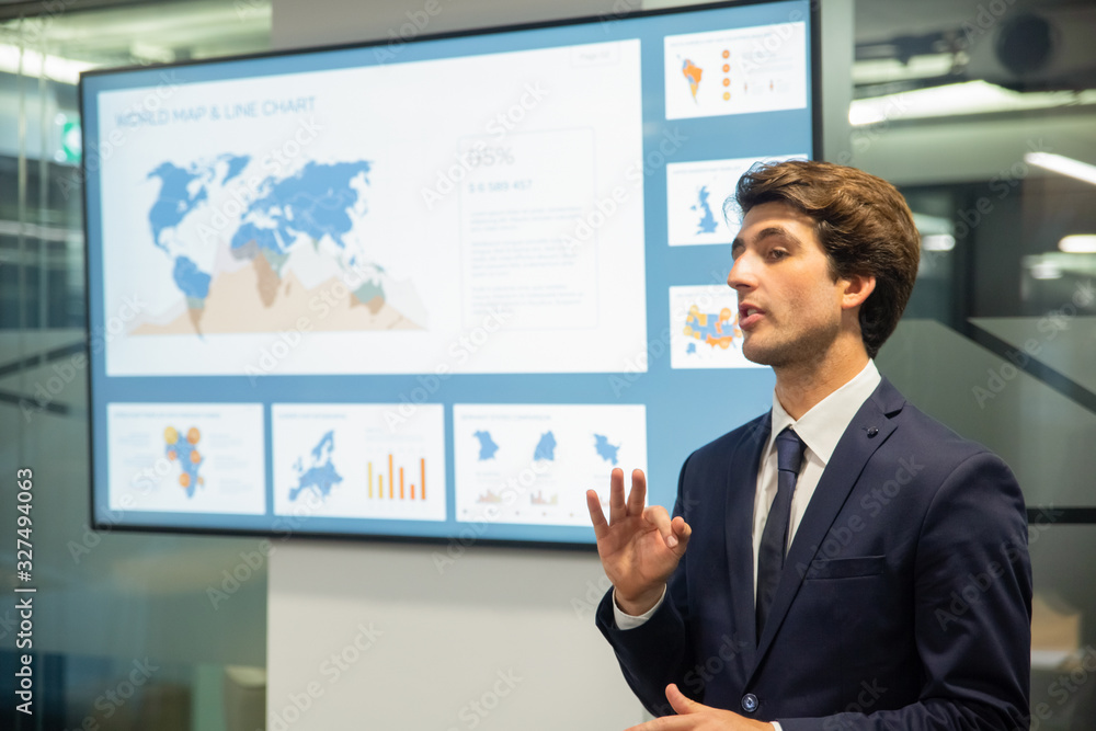 Confident man talking while standing near screen with information. Experienced analyst explaining details of new project. Business conference concept Confident man talking while standing near screen with information. Experienced analyst explaining details of new project. Business conference concept