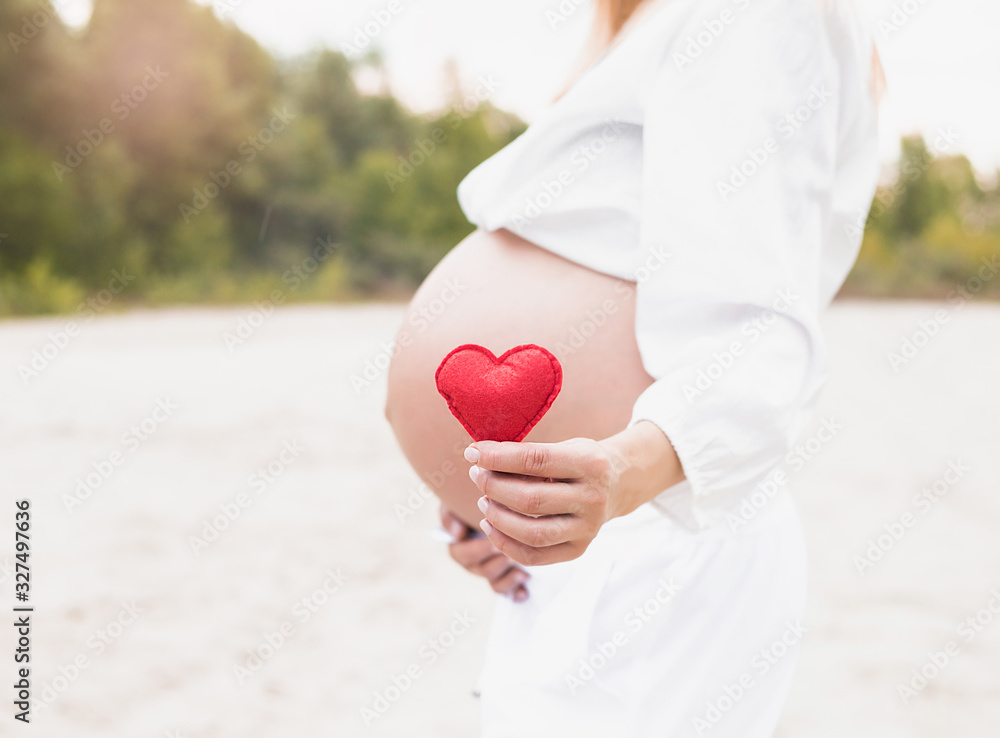 Young woman hand holding red bright heart shape on naked belly. Emotional loving moment in pregnancy time - 37 weeks. Baby expectation. Love, happiness and safety concept. Closeup. Young woman hand holding red bright heart shape on naked belly. Emotional loving moment in pregnancy time - 37 weeks. Baby expectation. Love, happiness and safety concept. Closeup.