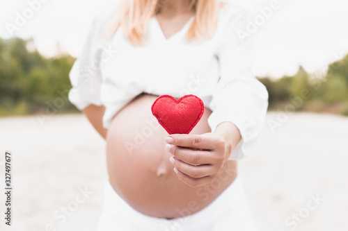 Young woman hand holding red bright heart shape on naked belly. Emotional loving moment in pregnancy time - 37 weeks. Baby expectation. Love, happiness and safety concept. Closeup.