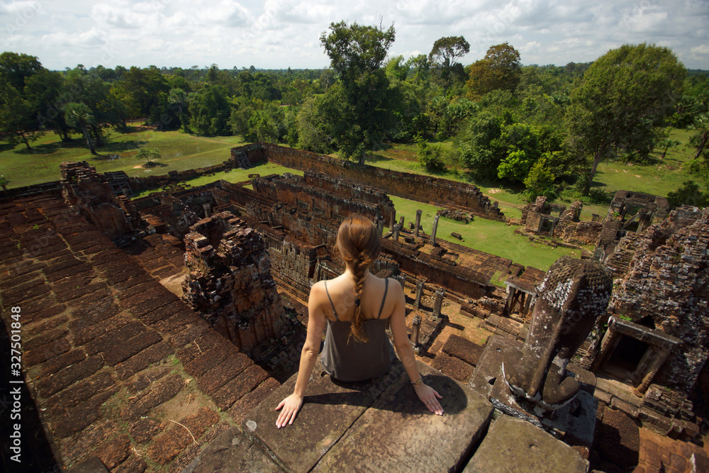 Fototapeta premium Young woman sitting on stones of old Angkor temple on background of bright green landscape and ancient ruins, Cambodia