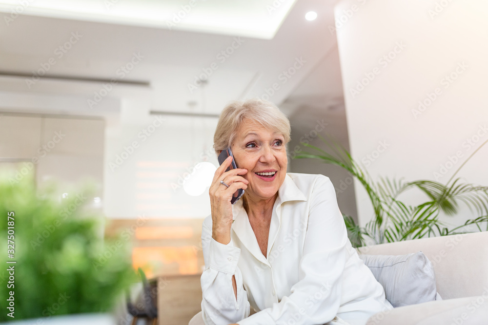 Senior woman talking on her mobile phone. Senior woman has a happy conversation at cellphone. Smiling senior woman using phone sitting on couch at home. Senior woman talking on her mobile phone. Senior woman has a happy conversation at cellphone. Smiling senior woman using phone sitting on couch at home.