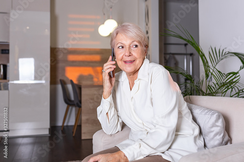 Happy senior woman using mobile phone at home. Smiling cool old woman with white hair sitting on sofa and messaging with smartphone. Beautiful stylish elderly lady talking on cellphone.