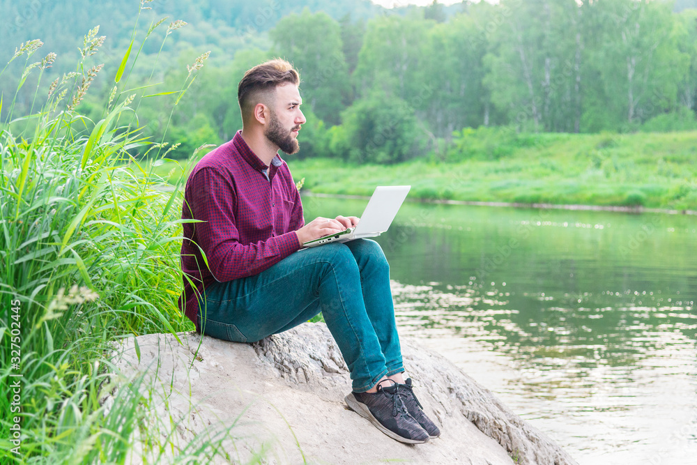 Fototapeta premium A young man in a shirt and jeans sits on a stone against the background of nature near the river on a summer day with a laptop. remote work and freelance concept
