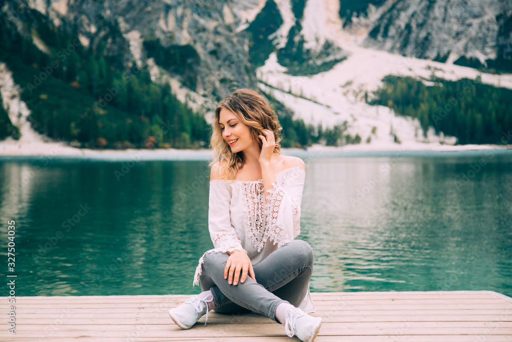 Beautiful girl long flowing hair sits on wooden pier, legs lotus position, hand near head smiles enjoy nature relax silence beauty of alpine Lake Braies. Backdrop white mountains green forest water