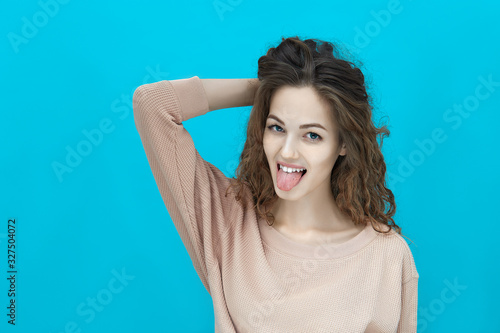 Portrait of a young beautiful woman wearing sweatshirt holds her hand on the back of her head shows tongue and looking at you isolated over blue background