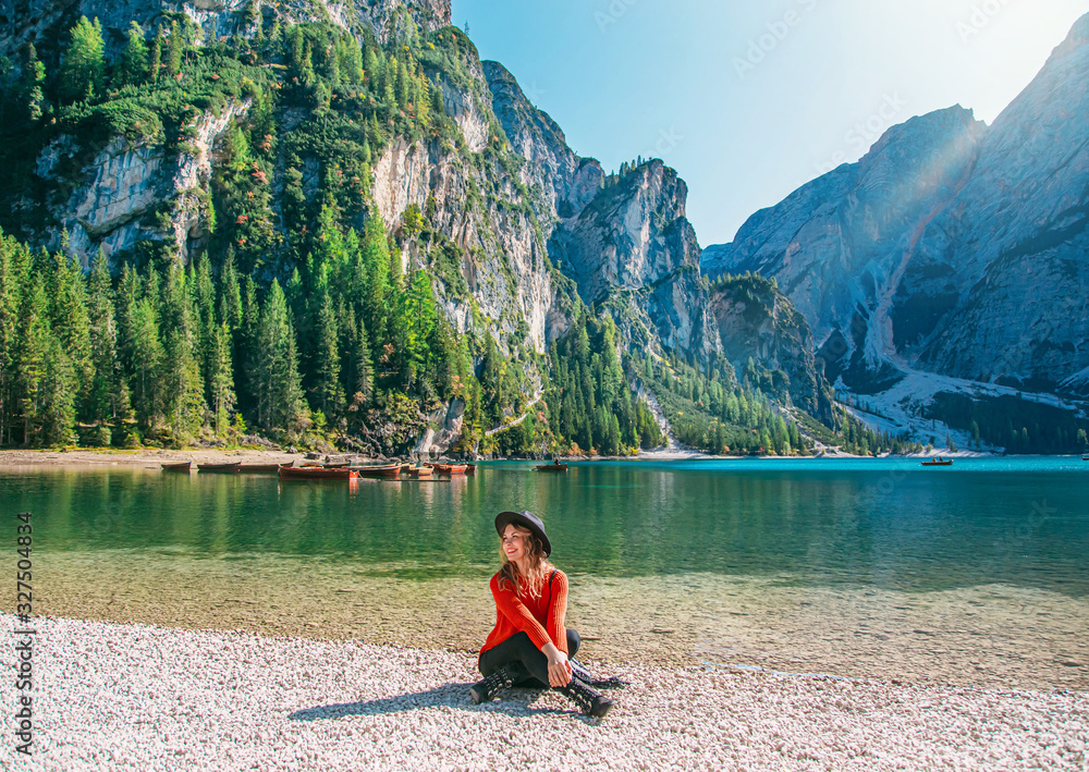 Вeautiful girl is resting on shores highland Lake Braies. Tourist smiles enjoy silence sunlight fantastic nature relax. Backdrop snowy mountains green forest reflected in water turquoise mint color
