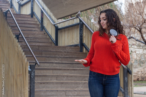 pretty caucasian young woman with magnifying glass copyspace imaginary on palm to insert advertisment, in the park, orange sweater and jeans, long curly hair. Place for your text in copy space.