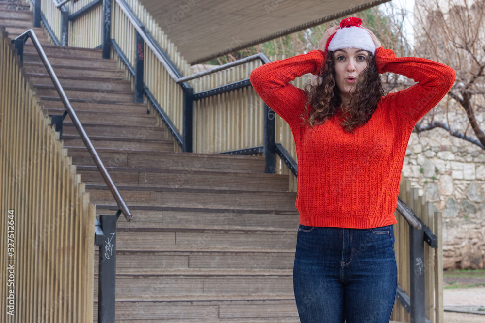 Portrait of attractive caucasian young woman with Santa Claus's hat being surprised, opening her eyes and mouth with shock, touching her head with hands, in the park, orange sweater and jeans, long cu