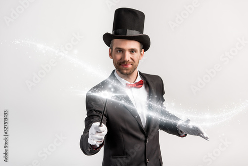 positive magician in suit and hat holding wand isolated on grey with glowing illustration