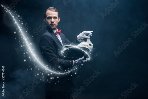 professional magician in suit showing trick with white rabbit in hat, dark room with smoke and glowing illustration