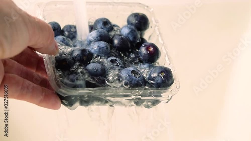 A woman's hand holds a plastic container with blueberries under the tap with water