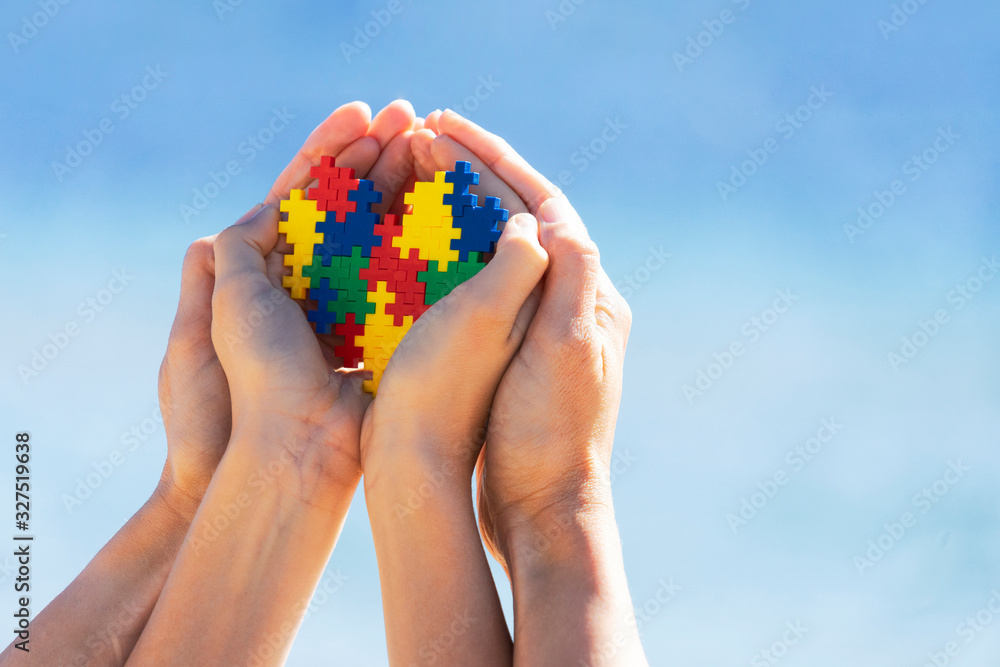 Child and adult hands holding colorful heart on blue sky background. World autism awareness day concept Child and adult hands holding colorful heart on blue sky background. World autism awareness day concept