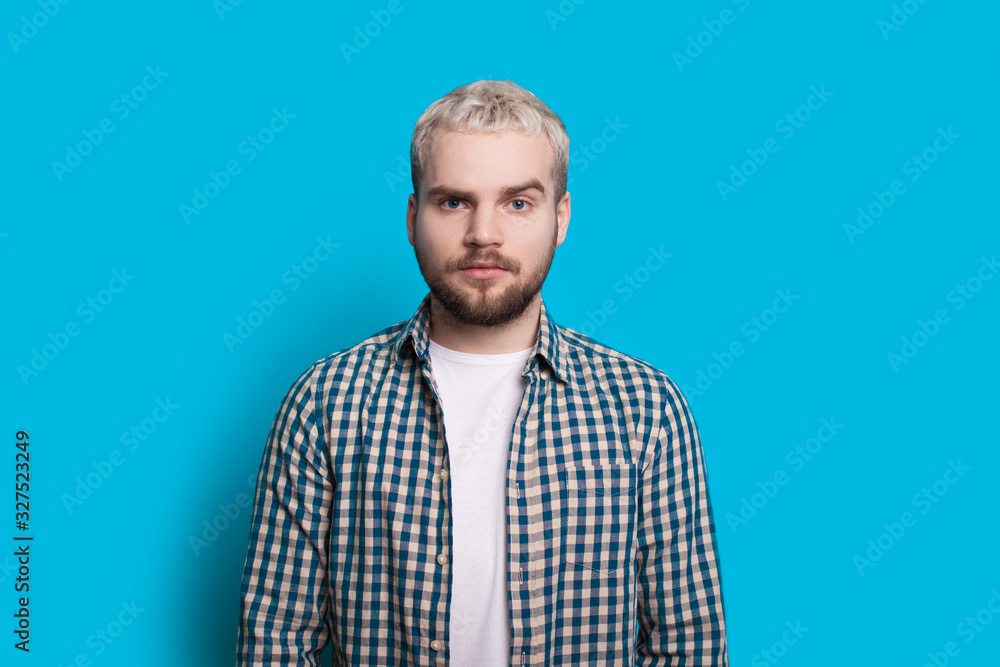 Obraz premium Caucasian man with blonde hair and beard is looking confidently at camera while posing on a blue wall