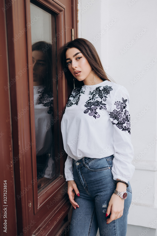 a young girl with brown hair in a white blouse and blue jeans poses against a light colored building a young girl with brown hair in a white blouse and blue jeans poses against a light colored building