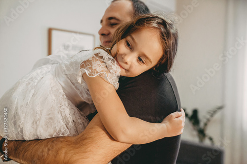 Charming caucasian girl dressed in a white dress looking back while her father is holding and embracing her