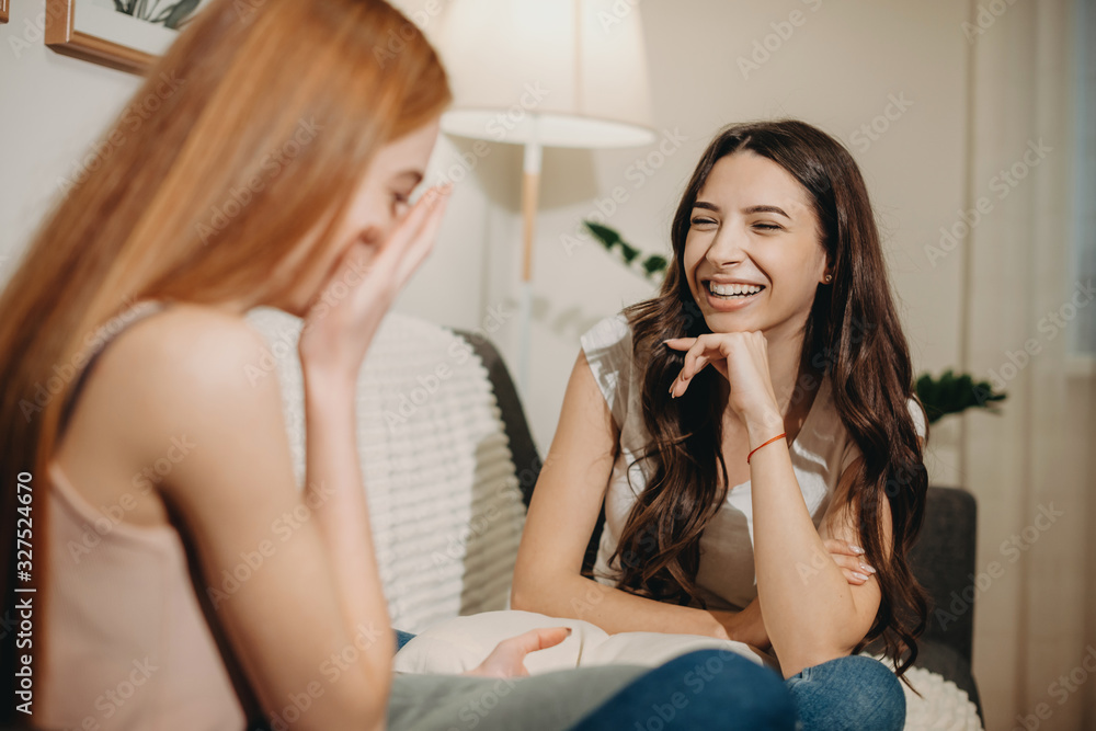 Funny ginger caucasian girl is telling a story to her brunette sister sitting together on the sofa and having joy Funny ginger caucasian girl is telling a story to her brunette sister sitting together on the sofa and having joy