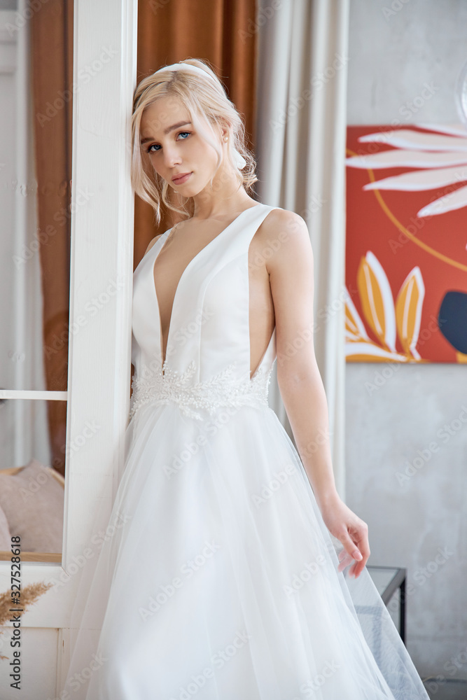 Beautiful slender blonde in the evening sun in a long white dress near a large window. Perfect hair and makeup for the bride, a new collection of wedding dresses
