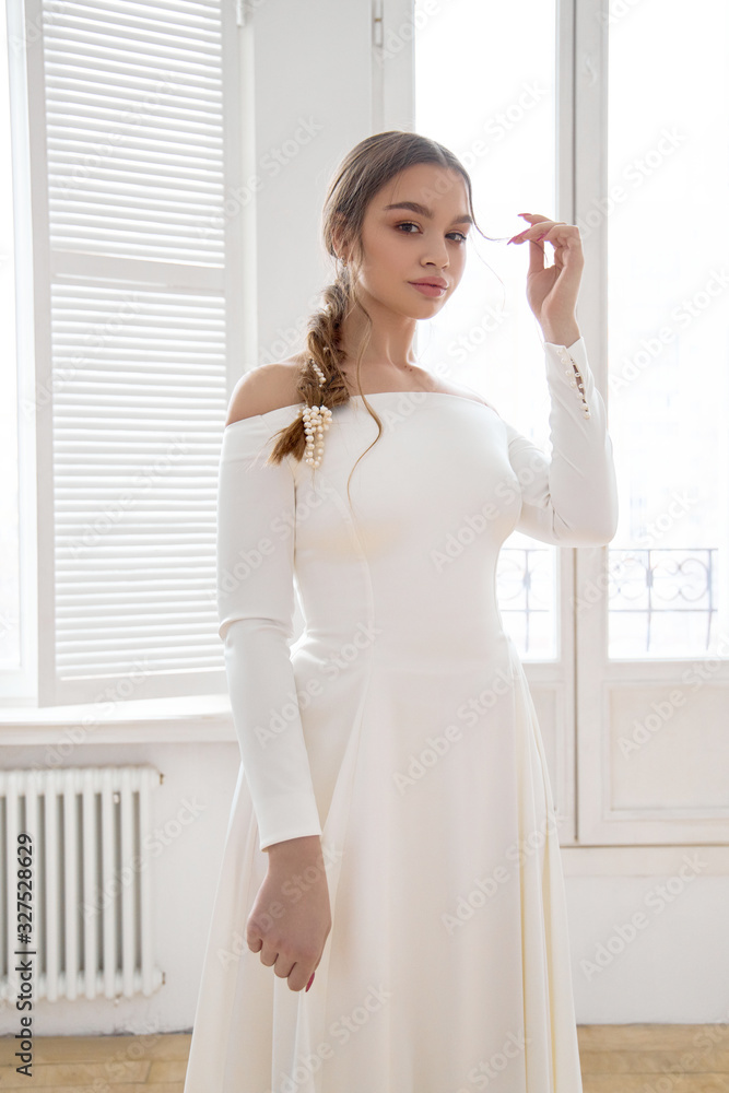 Beautiful slender blonde in the evening sun in a long white dress near a large window. Perfect hair and makeup for the bride, a new collection of wedding dresses Beautiful slender blonde in the evening sun in a long white dress near a large window. Perfect hair and makeup for the bride, a new collection of wedding dresses