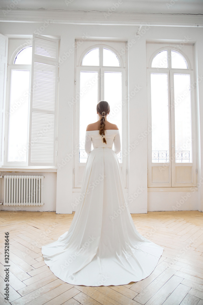 Beautiful slender blonde in the evening sun in a long white dress near a large window. Perfect hair and makeup for the bride a new collection of wedding dresses Stock Photo