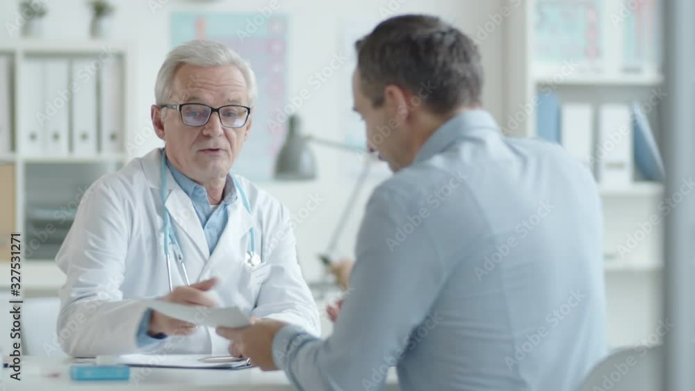 Senior doctor in lab coat shaking hands with young male patient, reading his medical history and giving consultation in clinic Senior doctor in lab coat shaking hands with young male patient, reading his medical history and giving consultation in clinic