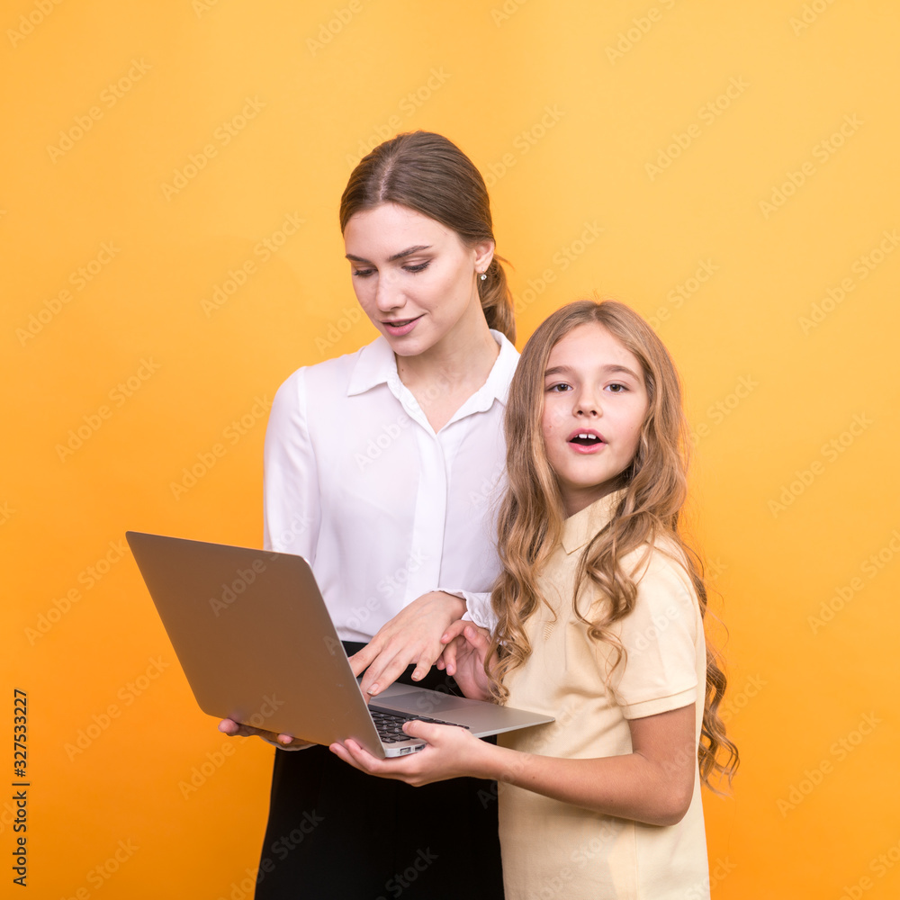 Beautiful teacher and cute schoolgirl hold in hands laptop for study on orange background. Beautiful teacher and cute schoolgirl hold in hands laptop for study on orange background.