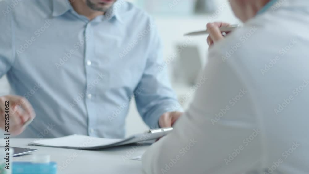 Tilt up of young Caucasian man taking clipboard from male doctor and signing medical service agreement during healthcare consultation in clinic Tilt up of young Caucasian man taking clipboard from male doctor and signing medical service agreement during healthcare consultation in clinic