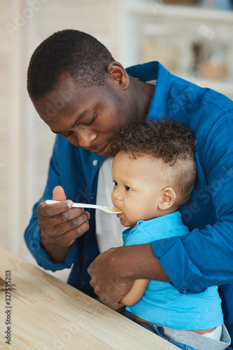 Side view portrait of happy African-American dad feeding cute little boy white sitting at kitchen table