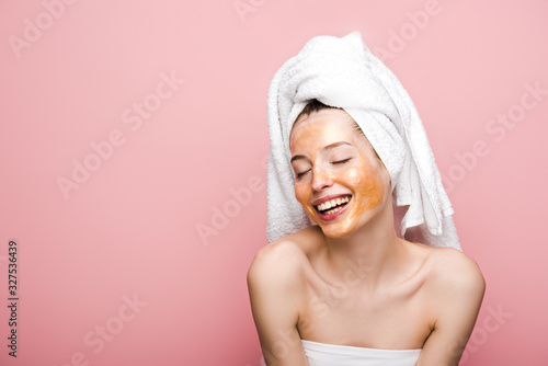 cheerful girl with facial mask and towel on head smiling with closed eyes isolated on pink
