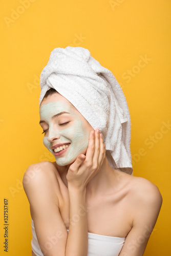 happy girl with nourishing facial mask and towel on head touching head with closed eyes isolated on yellow