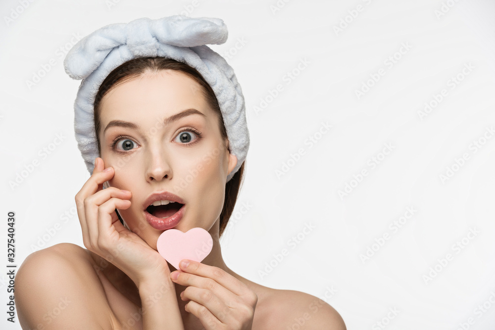shocked girl holding heart-shaped cosmetic sponge while looking at camera isolated on white shocked girl holding heart-shaped cosmetic sponge while looking at camera isolated on white