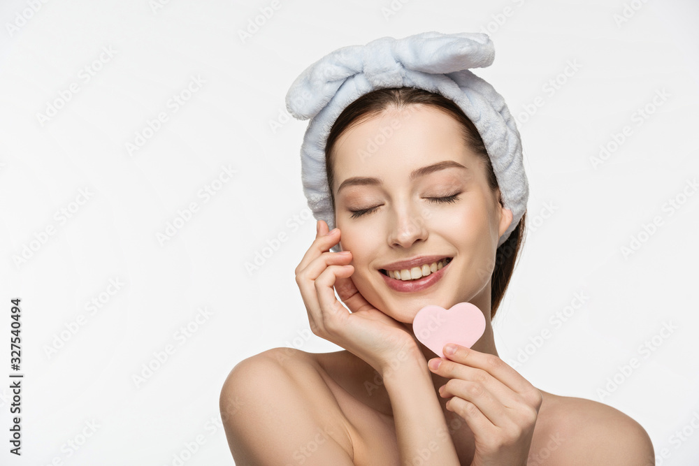 happy girl with closed eyes holding heart-shaped cosmetic sponge while touching face isolated on white happy girl with closed eyes holding heart-shaped cosmetic sponge while touching face isolated on white