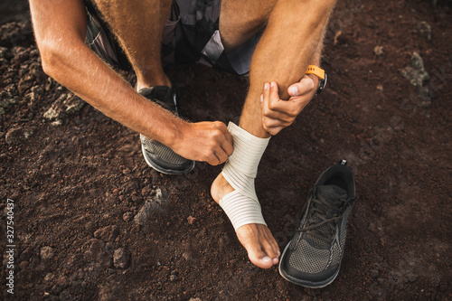 Man bandaging injured ankle. Injury leg while running outdoors. First aid for sprained ligament or tendon. Close-up on dark background.