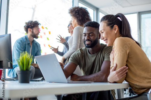 A man and a woman laughing while looking at the lap top with another man and a woman in the background of a workspace environment. 